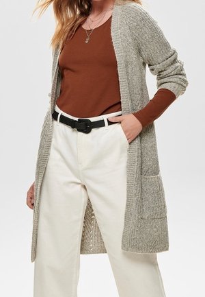 Femme portant un haut marron à manches longues, un cardigan en tricot beige avec des poches, un pantalon blanc taille haute et une ceinture noire, les mains dans les poches.