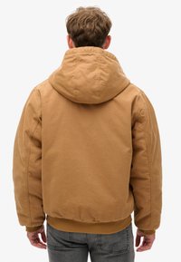 Chaqueta marrón con capucha, de textura suave, con puños y cinturilla elásticos, que presenta un corte holgado y un diseño minimalista.