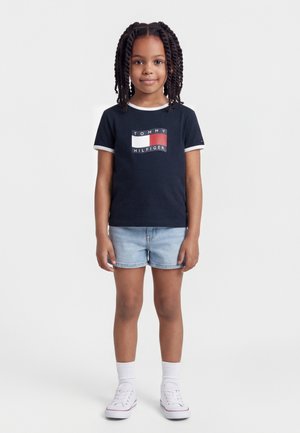 Ragazza giovane con capelli intrecciati, indossa una maglietta blu navy Tommy Hilfiger, pantaloncini di jeans chiari, calzini bianchi e scarpe da ginnastica bianche, in piedi su sfondo bianco.