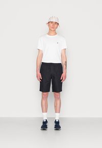 T-shirt bianco, pantaloni corti neri e sneakers blu navy. Tatuaggio visibile sul braccio, indossa un cappello a secchiello a righe. Aspetto atletico semplice e informale.