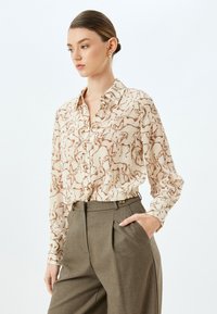 Beige blouse met een bruin paardenpatroon, knoopsluiting aan de voorkant en een kraag. Gecombineerd met grijze high-waisted broek met plooien aan de voorkant en zakken.