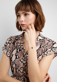 Gold-tone bracelet featuring a heart charm. The wearer has shoulder-length hair and is in a pink and brown snake print blouse.