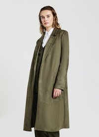 Cappotto lungo verde oliva con una texture liscia, rever a punta e tasche laterali, indossato sopra un completo abbinato e una camicia bianca.