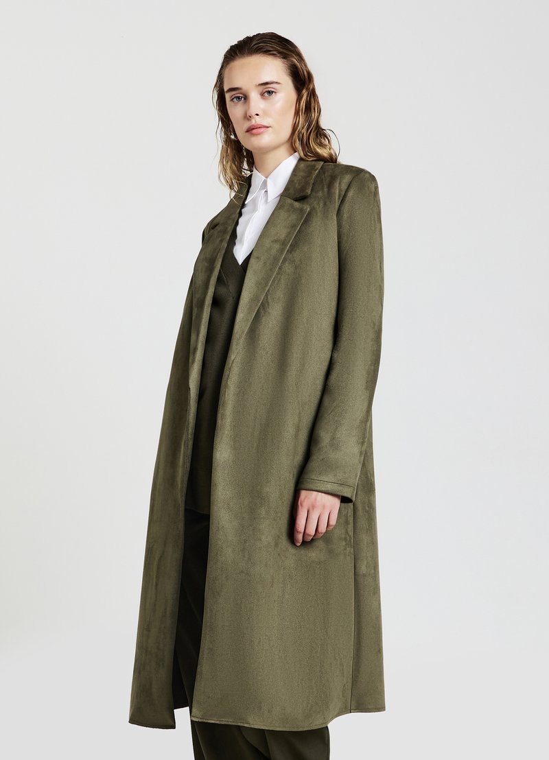 Cappotto lungo verde oliva con una texture liscia, rever a punta e tasche laterali, indossato sopra un completo abbinato e una camicia bianca.