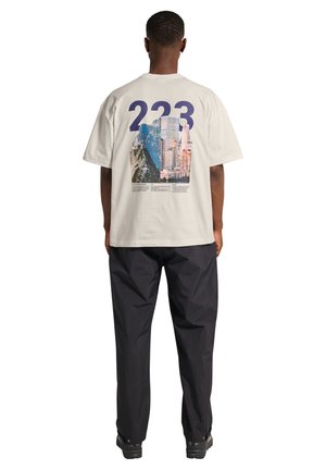 Mann trägt weißes T-Shirt mit großem "223"-Text und Stadtbild-Grafik auf der Rückseite, kombiniert mit lockeren schwarzen Hosen und schwarzen Schuhen.