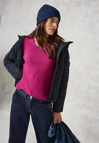 Marineblauer Puffer-Jacke über einem strukturierten fuchsiafarbenen Oberteil, kombiniert mit dunklen Jeans. Zubehör: gerippte marineblaue Mütze. Hält einen blauen Denim-Artikel.