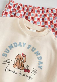 Cremfarbener Pullover mit der Aufschrift "SUNDAY FUNDAY" und einer Bunny-Grafik; rot und blau gemusterte Shorts mit elastischem Bund. Weicher Baumwollmix.