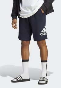 Námornícké modré bavlnené športové šortky s bielym logom Adidas, v kombinácii s sandálmi so čiernymi prúžkami a bielymi crew ponožkami. Neformálny a športový vzhľad.
