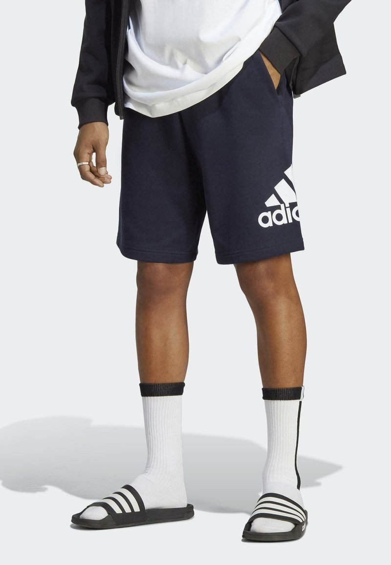 Námornícké modré bavlnené športové šortky s bielym logom Adidas, v kombinácii s sandálmi so �čiernymi prúžkami a bielymi crew ponožkami. Neformálny a športový vzhľad.