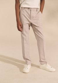 Pantaloni grigio chiaro con tessuto a trama strutturata, vestibilità sartoriale, pinces singoli e tasche laterali, abbinati a sneakers bianche.