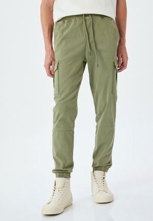 Groene cargobroek van zacht materiaal, voorzien van zijzakken, een trekkoord in de taille en elastische boorden. Draagt met lichtgekleurde sneakers.