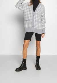 Cardigan grigio a motivo houndstooth con chiusura a bottoni, tasche frontali e polsini a costine, abbinato a pantaloni corti neri e stivaletti in pelle.