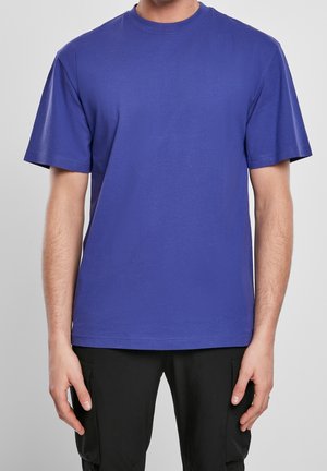 Uomo con maglietta blu tinta unita a maniche corte con scollo rotondo e pantaloni neri, in piedi con le braccia rilassate su uno sfondo grigio neutro.