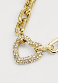 Bracciale in oro con charm a forma di cuore decorato con rhinestone trasparenti, con dettagli in metallo incisi con il nome del marchio "Guess."