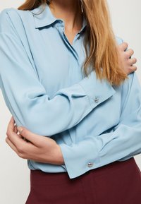 Světle modrá halenka s dlouhým rukávem, úzkým límcem a knoflíkovými manžetami, vyrobená z hladké látky a v uvolněném střihu, skombinovaná s tmavě burgundskými kalhotami.