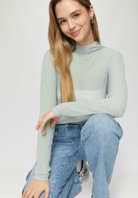 Lichtgroene geribbelde lange mouw top met een hoge hals, gecombineerd met blauwe high-waisted jeans. Zilverkleurige metallic schoenen zichtbaar.
