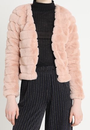 Personne portant une veste en peluche rose claire sur un col roulé noir côtelé et un pantalon rayé foncé.