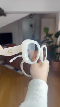 Balti per dydį dideli akiniai nuo saulės su apvaliais rėmeliais, skaidriais lęšiais ir auksine detalė ant rėmelio šono. Lygus plastikinis paviršius ir blizgus apdaila.