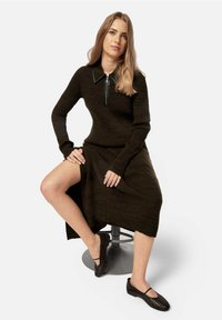 Rochie tricotată maro cu mâneci lungi și guler cu fermoar, având o fente pe lateral. Purtați cu pantofi țesuti negri, așezată pe un taburet argintiu.