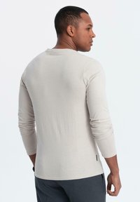 Hombre de espaldas vistiendo una camisa de manga larga color beige claro y pantalones gris oscuro, con un fondo blanco plano.