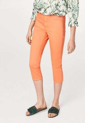 Person iført lyse orange cropped bukser kombineret med mørkegrønne sandaler med flæser og en grøn top med bladprint.