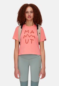 Różowy krótki t-shirt z logo „MAMMUT” w ciemnym brązie. Model ma na sobie jasnoniebieskie legginsy i beżowy plecak na ramieniu.