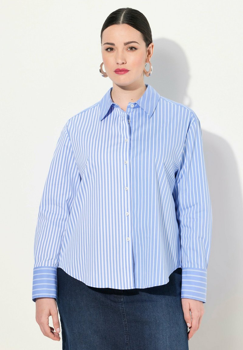 Donna con capelli scuri in una camicia a righe verticali blu e bianche con bottoni e una gonna scura, in piedi contro uno sfondo chiaro e semplice.