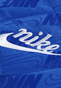 Kék steppelt szövet, dombornyomott Nike logókkal, a középpontjában egy feltűnő, fehér hímzett "Nike" swoosh logóval. Simogatós felület és tartós varrás.