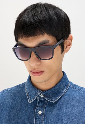 Uomo con capelli corti e neri indossa occhiali da sole rettangolari scuri e una camicia di jeans blu abbottonata su uno sfondo chiaro uniforme.