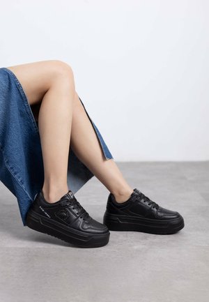 Las zapatillas de plataforma negras presentan cuero texturizado, un sistema de cordones y una suela de goma sólida. Se llevan con una falda de mezclilla que muestra una apertura lateral.