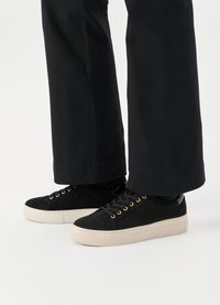 Schwarze Wildledersneaker mit goldenen Ösen und einer cremefarbenen Gummisohle. Merkmale sind eine abgerundete Zehenpartie und ein Schnürsystem, präsentiert mit schwarzen Hosen.