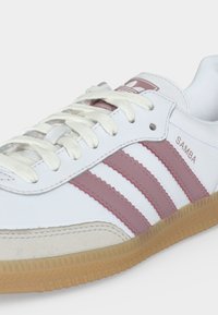 Λευκό αθλητικό παπούτσι Adidas Samba με ροζ ρίγες, λευκά κορδόνια, σόλα από καουτσούκ και εμφανές λογότυπο στο πλάι και στη γλώσσα.