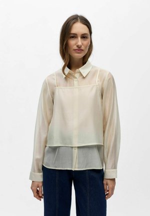 Kobieta o długich brązowych włosach, ubrana w przezroczystą kremową bluzkę nałożoną pod bezrękawowy kremowy krótki top oraz ciemnoniebieskie dżinsy, stojąca na białym tle.