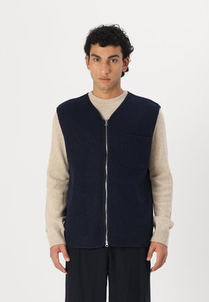 Navy wool vest with a high collar, zip front, and left chest pocket. Paired with a beige sweater featuring ribbed cuffs and round neck.