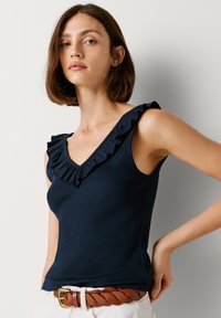 Marinblå ribbad linne med djup V-ringning och rynkad axeldetalj, kombinerad med ett flätat brunt skärp och ljusa byxor.