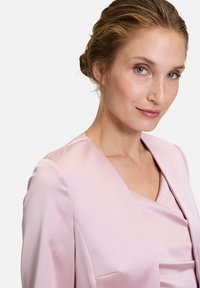 Vaaleanpunainen satiinitaakka, jossa on jämäkkä kaulus ja syvä V-pääntie. Pehme�ä tekstuuri vähäisellä yksityiskohtaisuudella, joka korostaa eleganttia ompelua.