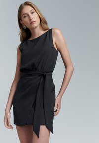 Frau mit langem blondem Haar, die ein ärmelloses schwarzes Wickelkleid trägt, das an der Taille gebunden ist, posiert vor einem schlichten grauen Hintergrund.