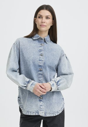 Lyseblå denimskjorte med knaplukning foran, klassisk krave og store ærmer. Har en let falmet tekstur og rundet kant.