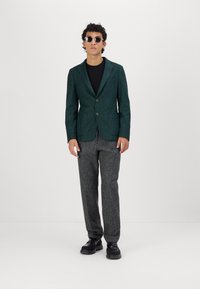 Mørkegrønn teksturert blazer med to knapper, kombinert med grå mønstrede bukser og svarte sko, brukt over en svart skjorte.