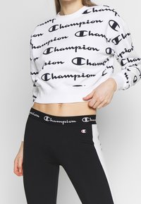 Kobieta ubrana w biały skrócony sweter i czarne legginsy, oba z powtarzającymi się czarno-białymi logo Champion.