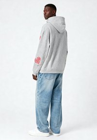 Sudadera gris con capucha y diseños de corazones rojos en las mangas, combinada con jeans holgados de color azul claro y zapatillas blancas. Textura de tela suave.