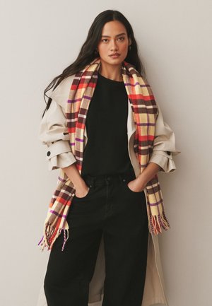 Hosszú sötét hajú nő bézs ballonkabátot, fekete felsőt és nadrágot visel, vörös, krémszínű és lila kockás sállal, kezét a zsebében.