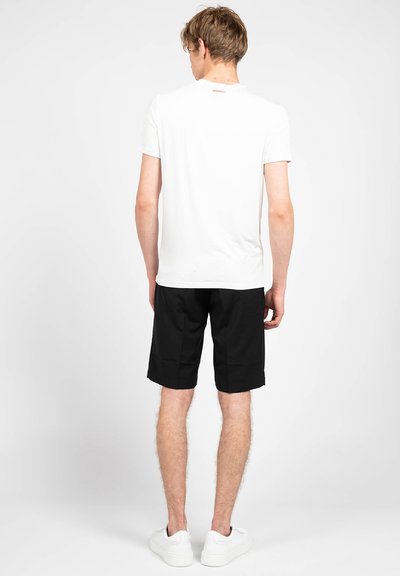T-shirt bianca in cotone con maniche corte, collo rotondo e logo discreto sul retro. Abbinata a pantaloni corti neri fino al ginocchio e sneakers bianche.