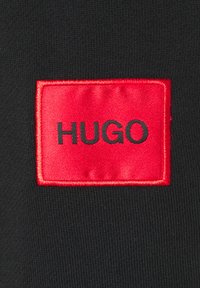 Rode rechthoekige stoffen patch met zwarte tekst "HUGO" op een achtergrond van zwarte gebreide stof genaaid.