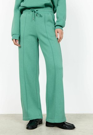 Person iført turkise vidde svedbukser med fremadrettede syninger og snøre i taljen, kombineret med sorte loafers, stående på et lyst gulv.