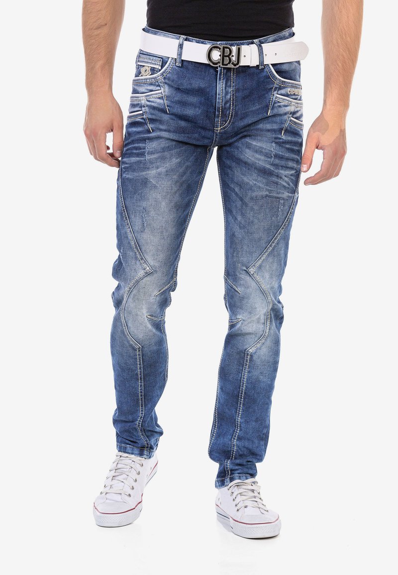 Blaue Jeans aus Denim mit einem ausgewaschenen Finish, ausgestattet mit detaillierten Nähten, fünf Taschen und einem weißen Gürtel mit Logo. Getragen mit weißen Sneakers.