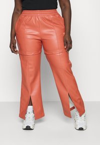 Coral faux leather pants with an elastic waistband, tapered fit, side slits, and snap details. Paired with white sneakers.