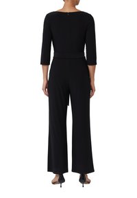 Tuta nera con un corpetto aderente, maniche tre quarti, una cerniera sul retro discreta e pantaloni a palazzo. Include una cintura coordinata in vita.