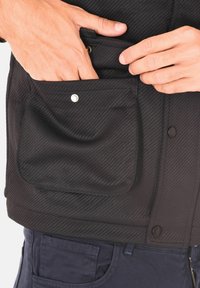 Giacca nera testurizzata con una tasca frontale visibile, chiusura a doppio bottone e dettaglio in tessuto a contrasto. Mano posizionata nella tasca.