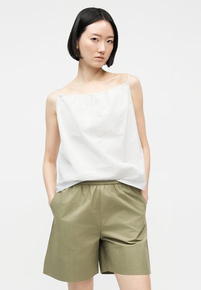 Γυναίκα με κοντά μαύρα μαλλιά, που φοράει λευκή ριγέ αμάνικη μπλούζα και χακί βερμούδα με ελαστική μέση, στέκεται με τα χέρια στις τσέπες.